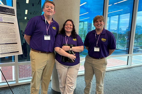 3 people wearing purple shirts with yellow lettering with the letters "GRPI" standing a smiling.