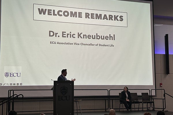 Speaker in front of projected powerpoint slide titled "Welcome Remarks, Dr. Eric Kneubuehl"