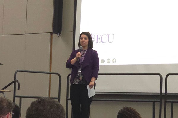 different angle of speaker with a microphone in one hand, and a piece of paper in the other. ECU logo being projected behind them.