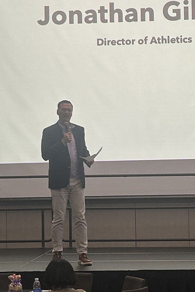Speaker holding a microphone and piece of paper. Projected in the background is "Jonathan Gil... Director of Athletics..."