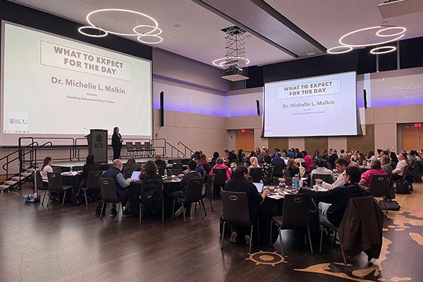 Slide being projected on 2 different screens in a room full of people. There is a speaker on stage. Tables full of people. The slide's title is "What to expect for the day by Dr Michelle L Malkin."