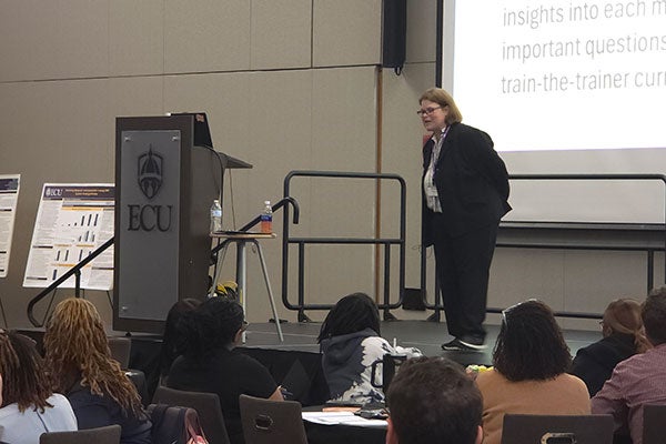 speaker on stage addressing someone in a crowd sitting at tables. Podium to left of speaker with ECU logo. Powerpoint slide being projected in the background.