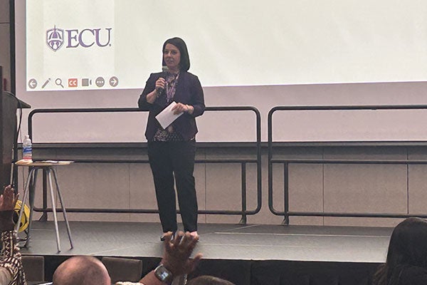 speaker with a microphone in one hand, and a piece of paper in the other. ECU logo being projected behind them.