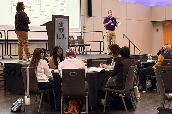 2 people standing on a stage. One man in a purple shirt is holding a microphone and a green piece of paper. Podium has ECU logo on it. Table in foreground with 6 people sitting. Another few people sitting to the right side of the stage.