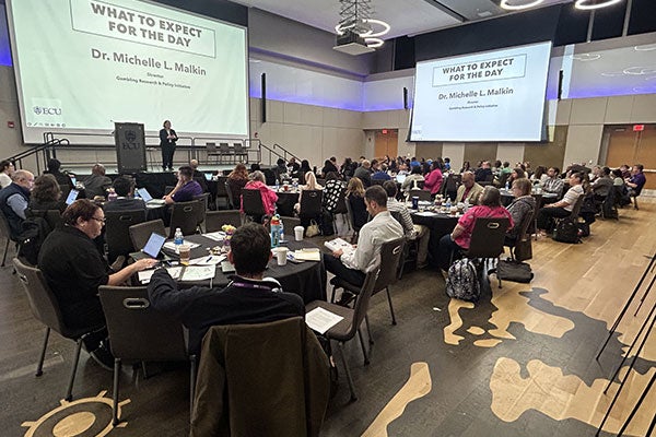 Different angle of Slide being projected on 2 different screens in a room full of people. There is a speaker on stage. Tables full of people. The slide's title is "What to expect for the day by Dr Michelle L Malkin."
